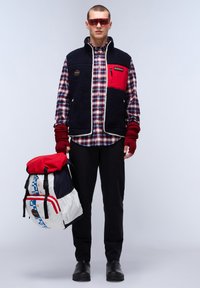 Marine fleece vest met rode borstpatch over een geruit shirt, zwarte broeken en zwarte schoenen; houdt een wit en rood gepatterne rugzak vast.