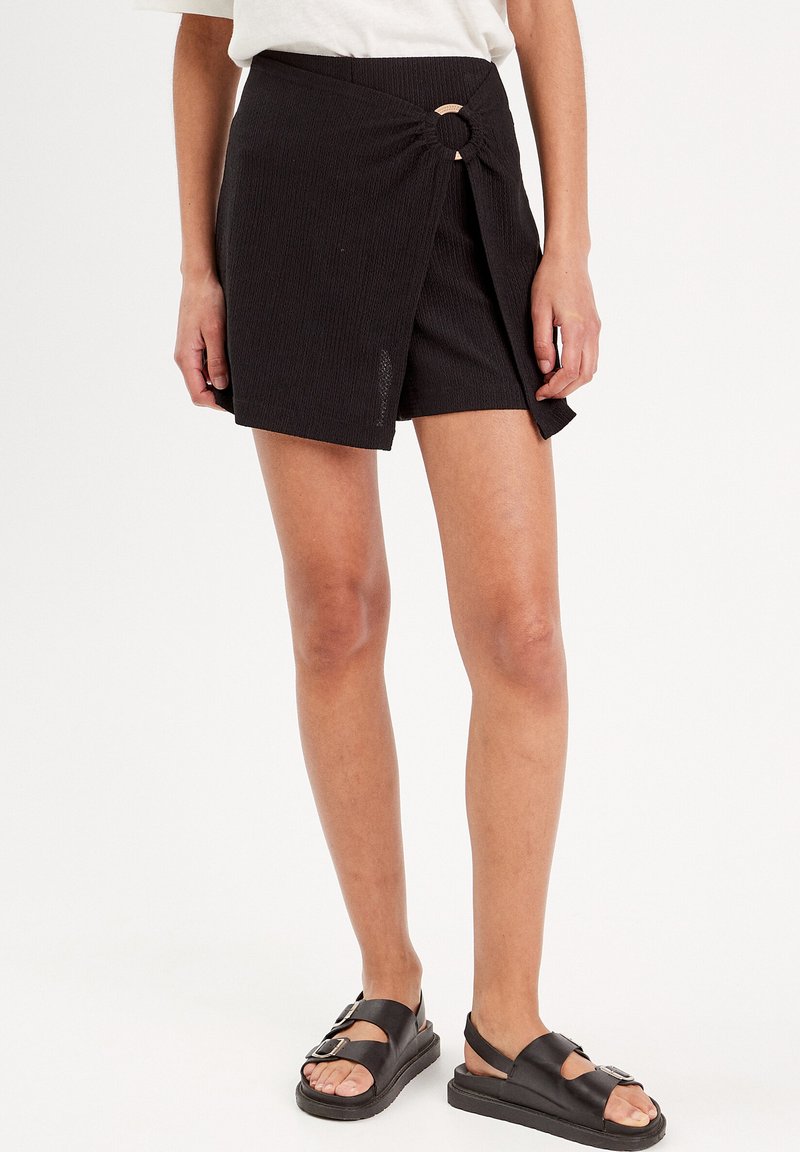 Shorts noirs de style enveloppant en tissu texturé, avec un accent en bois circulaire à la taille. À porter avec des sandales noires plates.