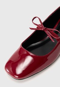 Sapatilha de balé de couro patente vermelho com bico quadrado e um laço correspondente na parte da frente. Textura suave e design simples.