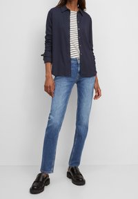 Persona con camisa azul marino abotonada sobre una blusa de rayas blancas y negras, jeans azules y mocasines de cuero negro, de pie contra una pared lisa.