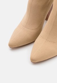 Beige pletené kotníkové boty s špičatou špičkou a texturovaným povrchem. Zvýšené podpatky a bezstehý design zvýrazňují moderní estetiku.