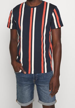 Man wearing navy blue t-shirt with vertical orange and white stripes and blue denim shorts with rolled cuffs, hand in pocket.