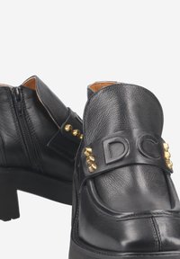 Schwarze Lederschuhe mit rundem Design, ausgestattet mit einem seitlichen Reißverschluss, einem geprägten "DC"-Riemen und goldenen Nieten als Akzente. Niedrige, klobige Absätze.