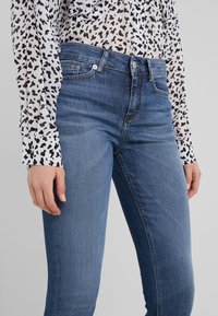 Vaqueros de mezclilla azul de corte slim, con un diseño clásico de cinco bolsillos y cierre de cremallera, combinados con una camisa blanca transparente con manchas negras.