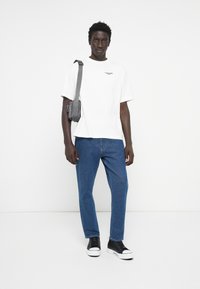 White cotton t-shirt with logo, blue jeans, and black sneakers. Grey crossbody bag on shoulder. Simple design, straight cut.