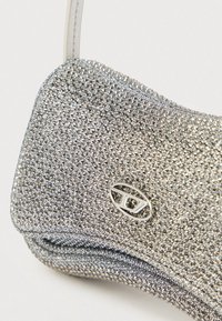 Geantă de mână argintie cu o suprafață texturată acoperită cu mici rhinestones. Prezintă un logo metalic și o formă curbată și elegantă.