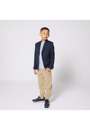 Marineblazer, lichtblauw overhemd, beige broek en zwarte sneakers. Blazer heeft één borstzak en twee zijzakken. Casual uitstraling.