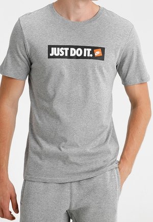 Homme portant un t-shirt gris Nike et un pantalon de survêtement, le t-shirt porte l'inscription "JUST DO IT." avec le logo Nike en orange et blanc.