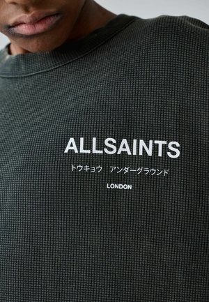 Nahaufnahme eines dunklen, strukturierten Hemdes mit weißem Text "ALLSAINTS", japanischen Schriftzeichen und darunter "LONDON".