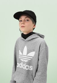 Sweat à capuche gris avec le logo Adidas blanc et trois bandes, assorti d'une casquette noire et d'un bandeau en mesh noir. L'arrière-plan est vert clair.