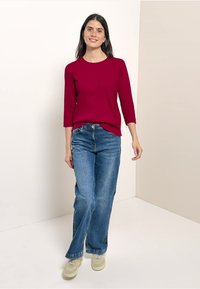 Rotes Oberteil mit drei Viertelärmeln, kombiniert mit hellblauen weiten Jeans. Das Modell trägt beigen Sneakers. Einfaches Design, glatte Textur und lässige Passform.