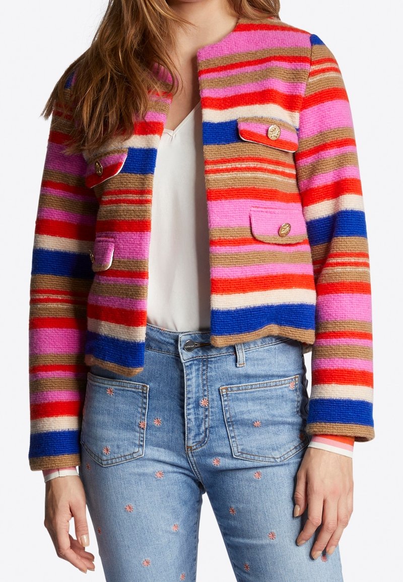 Femme portant une veste rayée vibrante avec des couleurs rose, rouge, bleu et beige sur un haut blanc, associée à un jean bleu clair brodé de petites fleurs roses.