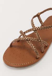 Sandale en cuir marron avec des brides en bronze métallisé tressées et de petites perforations sur le confort, présentée sur fond blanc.