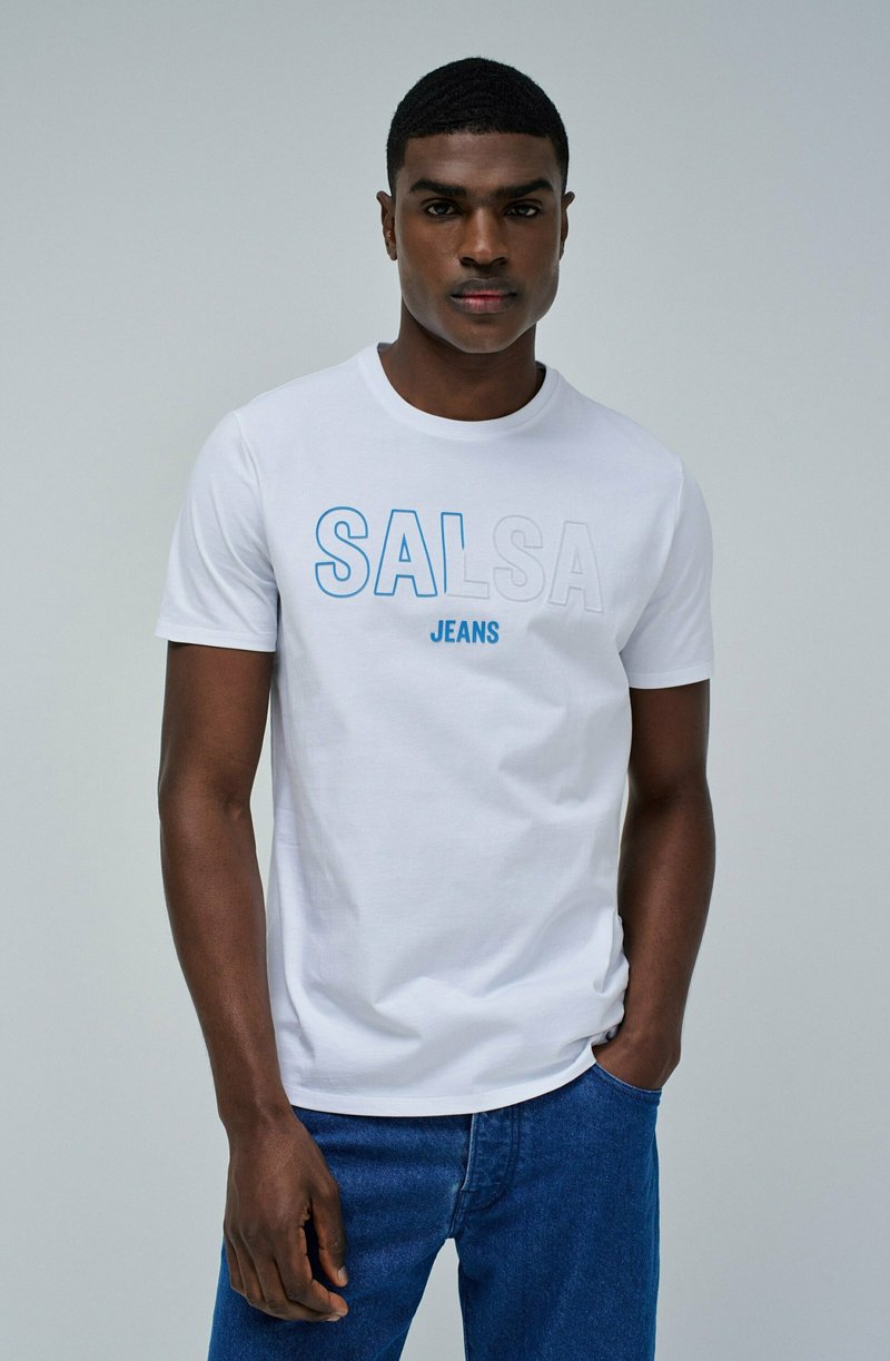 Camiseta blanca de cuello redondo con el texto "SALSA" en azul y gris claro, combinada con jeans de denim azul. Tejido de algodón suave.