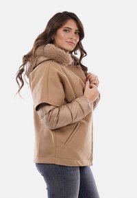 Cappotto beige in misto lana con maniche imbottite e cappuccio foderato in pelliccia. Presenta tasche laterali e chiusura a zip, caratterizzato da un design moderno e stratificato.
