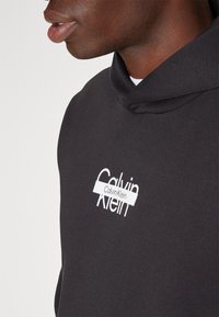 Felpa nera in morbido cotone con tasca a marsupio, caratterizzata da un logo bianco di Calvin Klein sul davanti e un cappuccio con cordino.