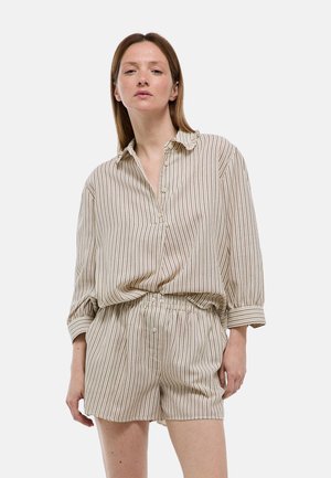 Femme portant une chemise à manches longues beige et marron rayée verticalement avec col et short assorti à taille élastique, debout devant un fond blanc.