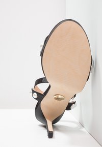 Czarna szpilka z perłowymi detalami, beżową podeszwą i złotym okrągłym logo na łuku, umieszczona podeszwą do góry na białym tle.