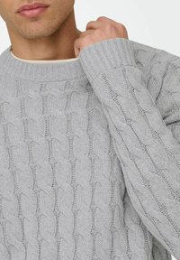 Homme ajustant le col d'un pull en tricot torsadé gris clair, montrant le motif de tricot texturé et les poignets et le col côtelés.