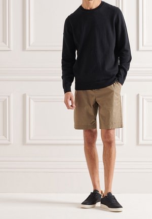 Homme portant un pull noir à manches longues, un short beige arrivant aux genoux et des baskets noires, debout contre un mur à panneaux blancs.