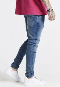 Jean ajustado en denim azul claro, con bolsillos traseros y un acabado desgastado, combinado con zapatillas blancas de corte bajo.