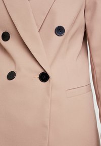 Blazer beige de doble botonadura con tela texturizada, presenta botones negros y un bolsillo lateral. Corte a medida y diseño estructurado.