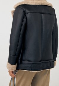 Veste en cuir noir avec un col et des poignets en peau lainée beige. Le dos présente des détails cousus et une finition lisse.