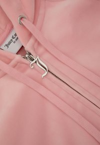 Roze zip-up hoodie gemaakt van zachte stof, met een zilveren rits met een decoratief ontwerp en bijpassende trekkoorden voor de capuchon.