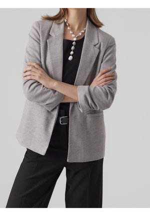 Person mit verschränkten Armen, trägt einen grauen Blazer, ein schwarzes Oberteil, schwarze Hosen, einen silbernen Gürtel und eine auffällige silberne Halskette.