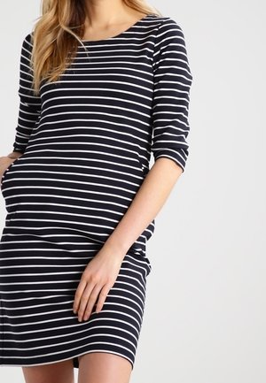 Mujer con un vestido de rayas horizontales en blanco y negro y mangas tres cuartos, de pie contra un fondo claro y liso.