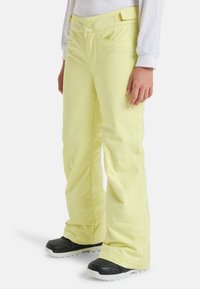 Gele sneeuwbroek met een recht gesneden design, voorzien van riemlussen, voorzakken en versterkte manchetten, gecombineerd met zwarte sneeuwlaarzen.