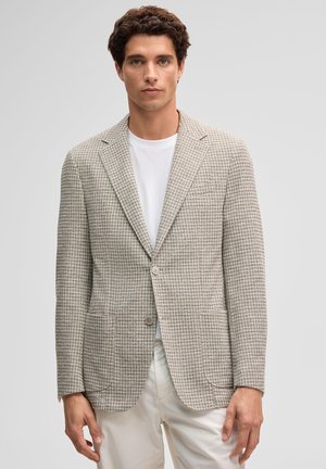 Giovane uomo che indossa un blazer beige a fantasia pied de poule sopra una t-shirt bianca con pantaloni color crema, in piedi su uno sfondo grigio chiaro uniforme.