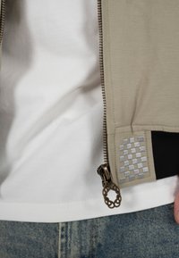 Gros plan sur la fermeture éclair d'une veste beige avec un tirette décoratif en bronze, broderie argentée à carreaux, portée sur une chemise blanche et un jean bleu.