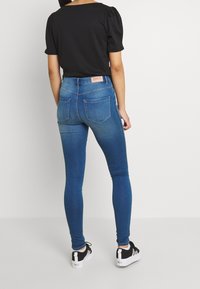 Vaqueros ajustados de color azul con cintura alta, un desvanecimiento sutil y una etiqueta de marca rosa en la parte trasera. Combinados con una camiseta negra de mangas abullonadas y zapatillas negras.