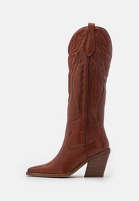 Bota de vaquero de cuero marrón con caña alta, puntera puntiaguda y costuras decorativas. Cuenta con un tacón apilado y detalles texturizados.