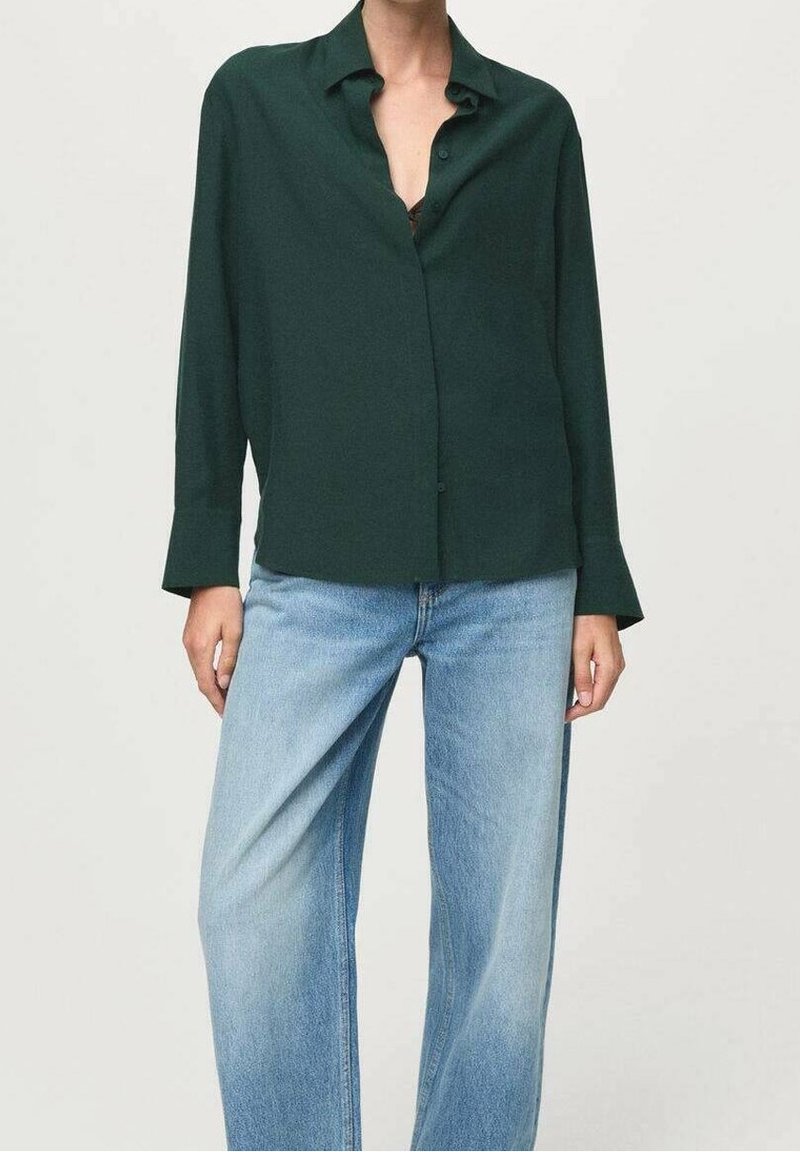 Personne portant une chemise boutonnée à manches longues vert foncé associée à un jean taille haute à jambes larges bleu clair sur un fond uni.