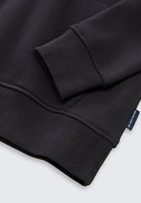 Sudadera negra con puños y dobladillo acanalados. Presenta costuras a juego y una etiqueta de tela en el costado. Material suave y texturizado.