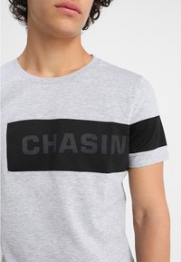 Mladý člověk v světle šedém tričku s širokým černým vodorovným pruhem a jemným textem "CHASIN" přes hruď.