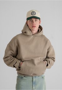 Beige oversized hettegenser med lomme foran, ribbestrikkede mansjetter, brukt med lyseblå jeans. Komplementert av en kremfarget caps med grønn skygge og logo.