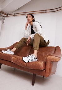 Zapatillas deportivas blancas y negras, pantalones verde oliva ajustados y un cárdigan gris claro sobre una camiseta negra, sentado en un sofá de cuero marrón.