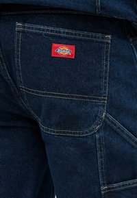 Σκούρο μπλε παντελόνι από denim με κίτρινα ραφές και κόκκινη ετικέτα λογότυπου, με πίσω τσέπη και καμπυλωτό σχεδιασμό τσέπης.