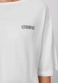 Camiseta de algodón blanca con escote redondo y mangas cortas. Presenta un pequeño logo impreso en negro "STUDIO45" en la parte superior izquierda.
