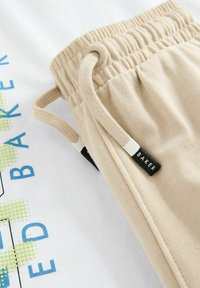 Beige snorebukser med elastisk talje og et sort mærke med "BAKER." Baggrunden viser en hvid skjorte med blå og grønne grafik.