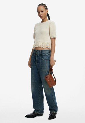 Top corto beige lavorato a maglia con orlo frangiato, abbinato a jeans blu larghi. Borsa marrone con dettagli di borchie. Stivali neri in stile western.