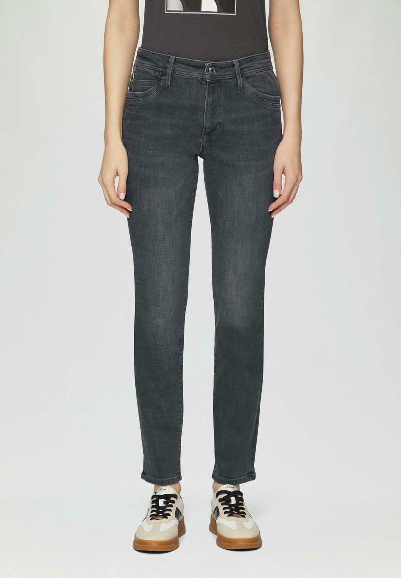 Jean en denim gris foncé à coupe slim, avec des détails de poches discrets et des ourlets légèrement effilochés, associés à des baskets beige et noires.