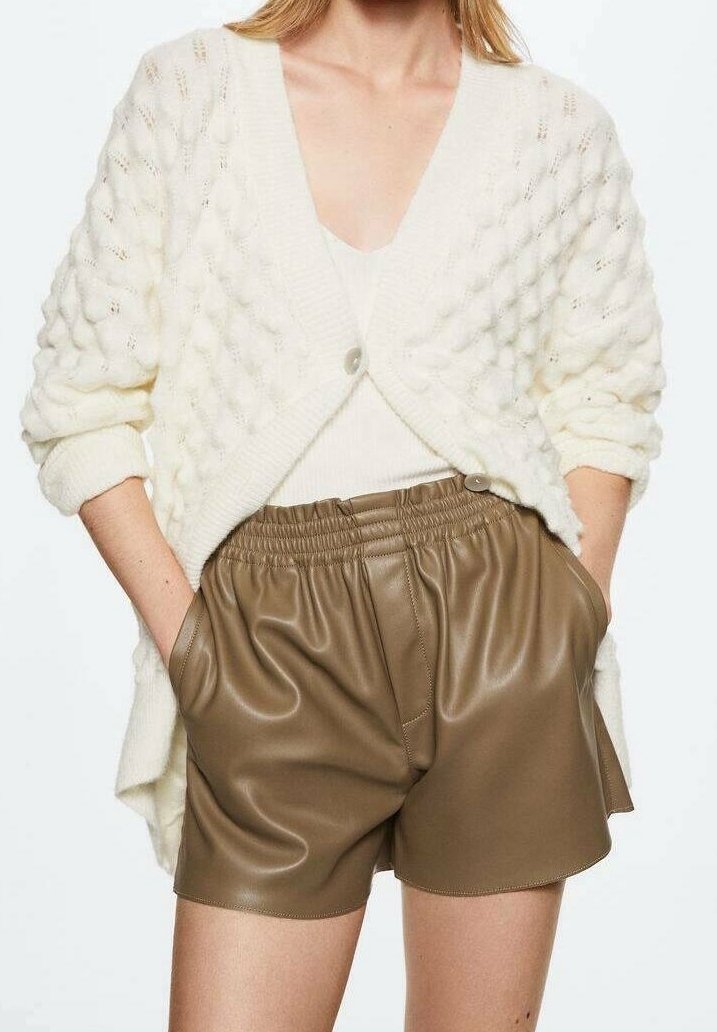 Femme portant un cardigan en maille texturée blanche sur un haut blanc, associé à un short en cuir marron taille haute avec une ceinture élastique et des poches.