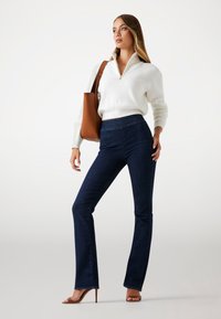 Jean flare bleu foncé en denim associés à un pull en tricot blanc texturé avec un col zippé et un sac à main en cuir marron.