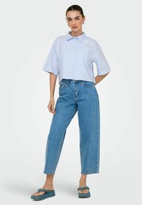 Camisa de rayas azul y blanca, recortada, con cierre de botones y mangas cortas, combinada con un pantalón de mezclilla claro de tiro alto y sandalias verdes.