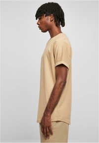 Tricou din bumbac bej cu mânecă scurtă și guler rotund, având o croială relaxată și un tiv rotunjit, purtat cu pantaloni bej asortați.