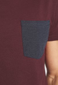 Detailní záběr na vínové tričko s tmavě šedou kapsou na hrudi, umístěné na horní části trupu a paži osoby.